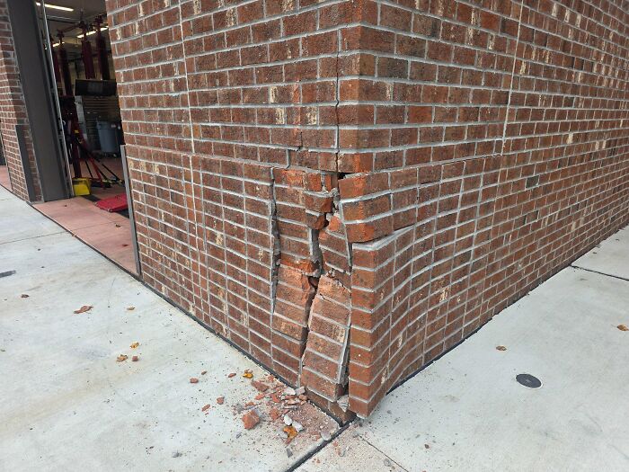 Car mechanics observe a damaged brick wall corner, with bricks dislodged and crumbling at the base of a building.