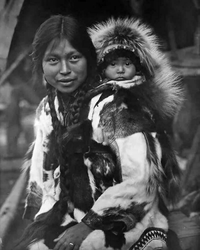 A historical photo of an indigenous woman in a fur coat with a baby in a fur-lined carrier on her back.