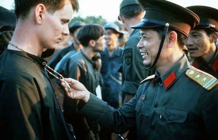 A historical photo showing a US soldier being greeted by a Vietnamese officer, exchanging a necklace.