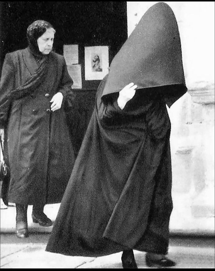Two women in dark, traditional clothing, one with a large conical hood obscuring her face, creating a mysterious and captivating historical photo.