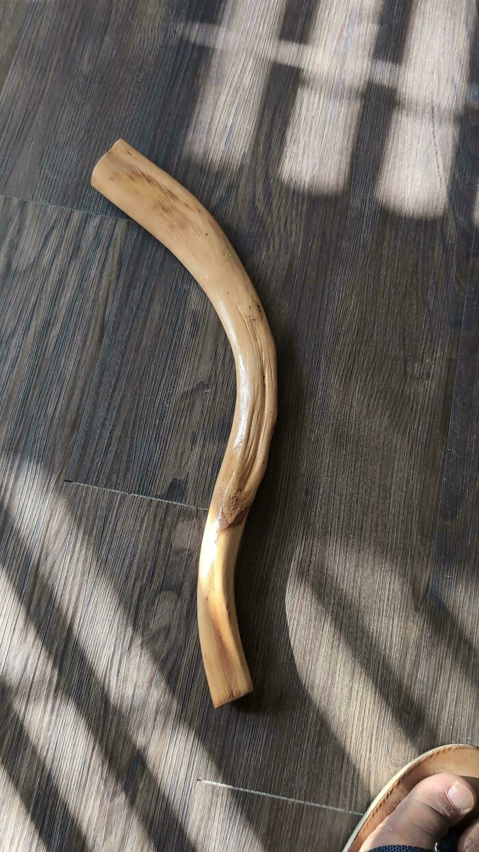 Curved wooden stick resting on a dark floor with light and shadow patterns near a person's foot nearby.