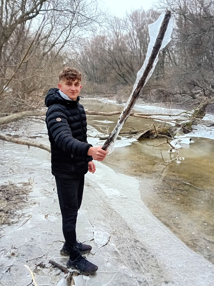 Young person outdoors holding a cool stick covered in ice near a frozen river in a winter forest setting.