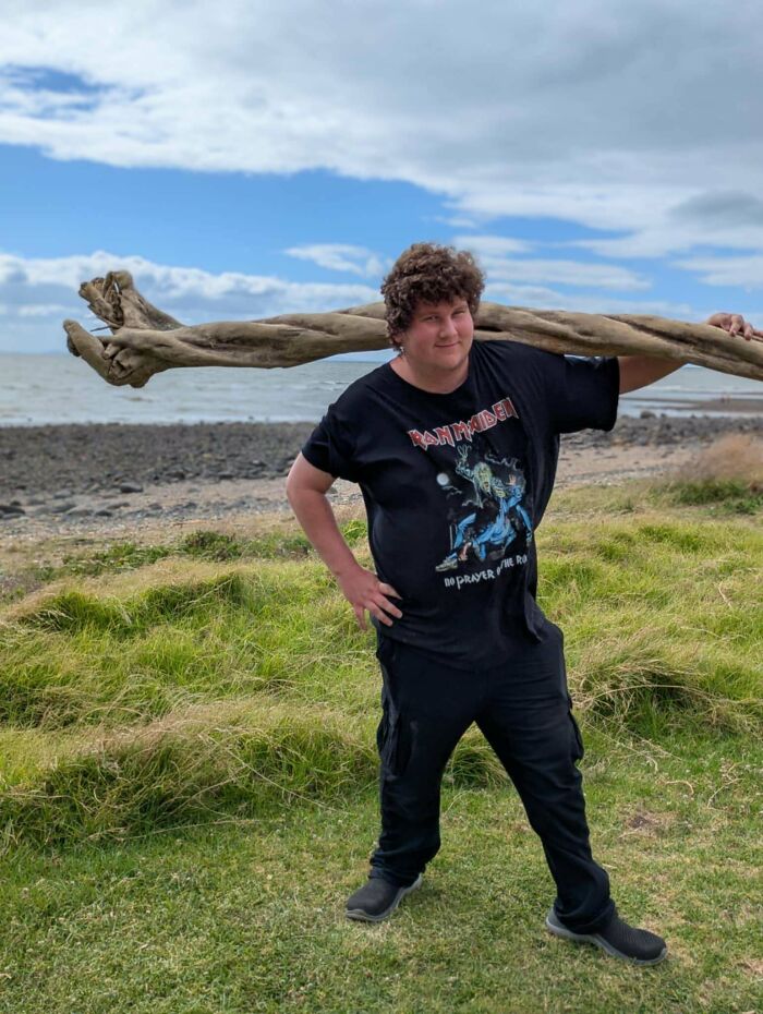 Person standing on grassy shore carrying cool sticks over their shoulders near the ocean under a cloudy sky.