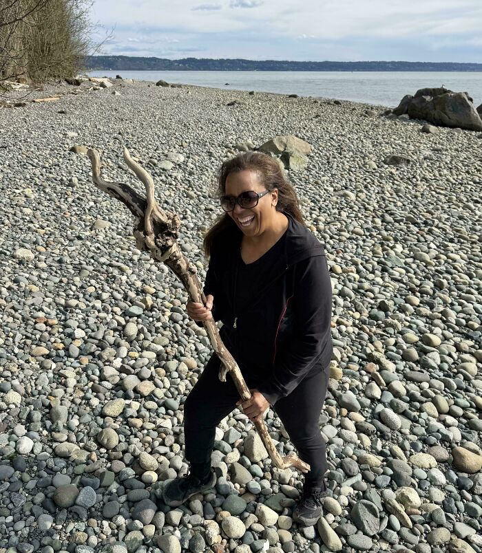 Woman sharing cool sticks on a rocky beach, smiling and holding a large twisted driftwood under cloudy sky.