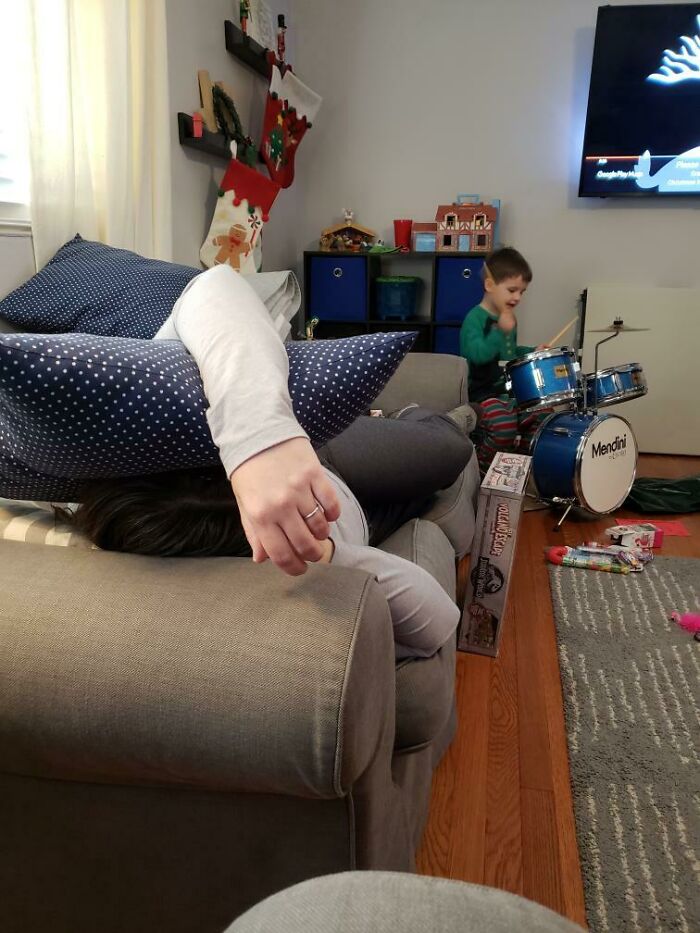 Adult hiding under pillows on a couch while a child plays drums, a funny regret moment in a family living room.