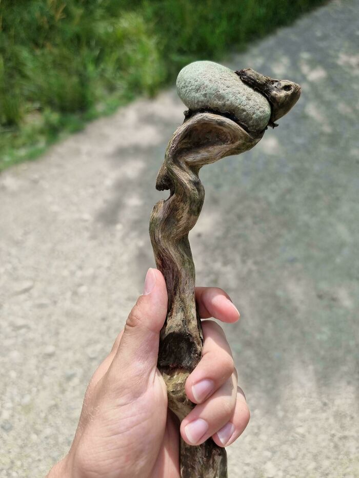 Hand holding a cool twisted wooden stick with a stone attached, displayed outdoors on a sunny day.