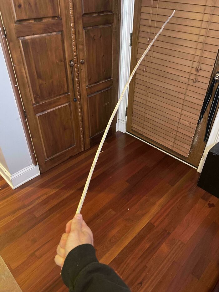 Hand holding a long, thin stick indoors with wooden floors and cabinets visible in the background.