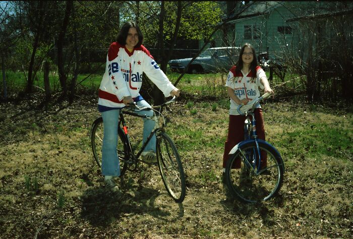 Two people wearing matching hockey jerseys on bicycles in a backyard capturing an awkward childhood moment.