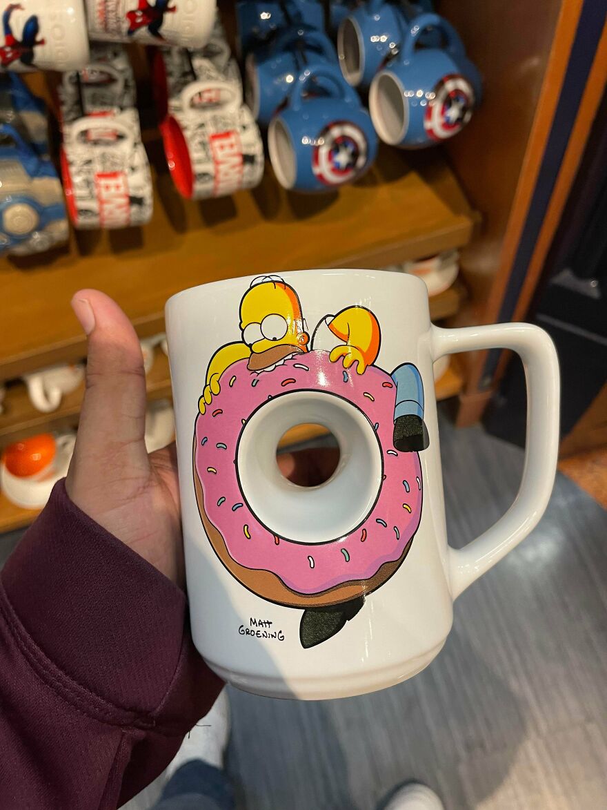 Hand holding an impossible to use mug featuring Homer Simpson with a donut-shaped hole in the center, showcasing flawed design.