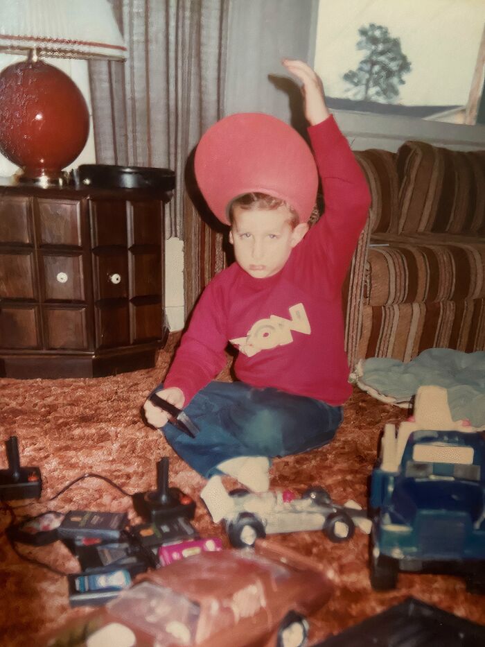 Child playing with toy cars and remote controls on carpet, wearing a red sweatshirt and a large red hat in an awkward childhood moment.