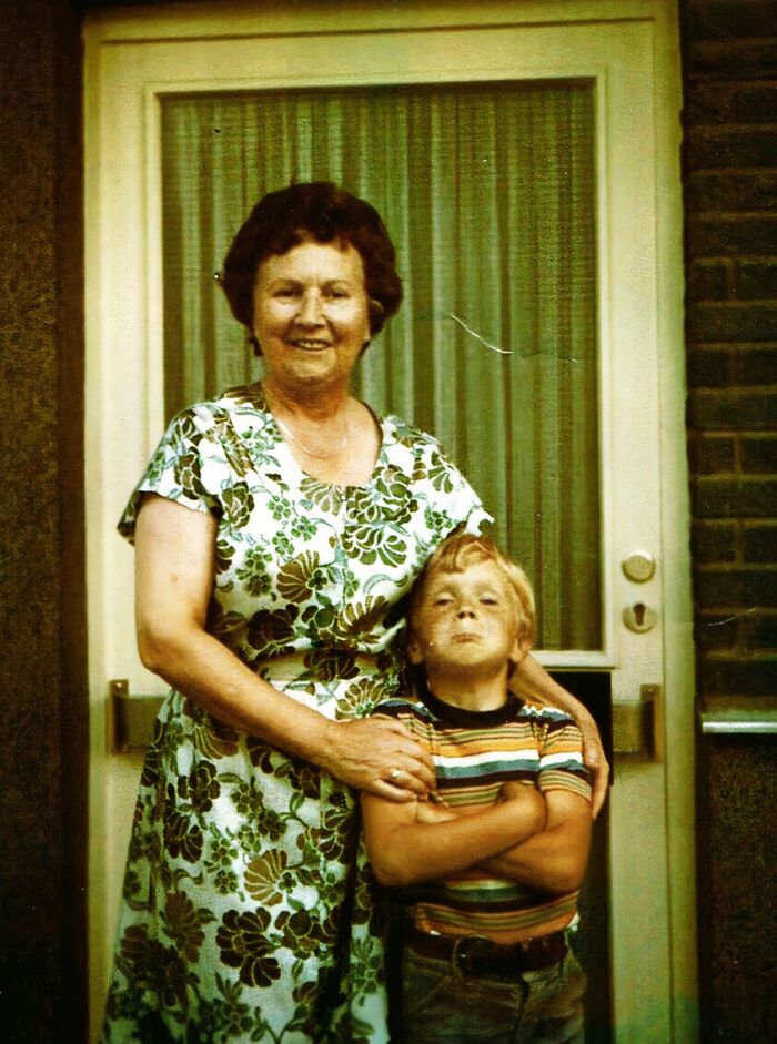 Vintage awkward childhood photo of a boy with crossed arms standing next to a smiling woman outside a door.