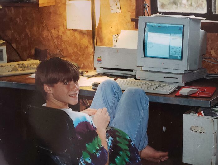 Child smiling awkwardly while sitting barefoot in front of a vintage computer, a classic childhood photo with nostalgic vibes.