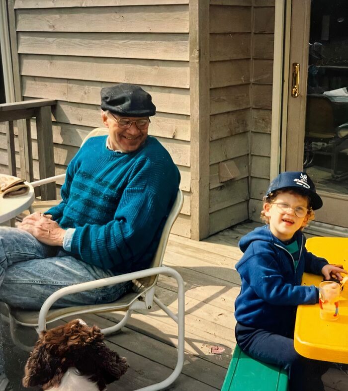 Elderly man and child wearing hats, sitting outside on a deck with a dog nearby in a childhood awkward photo moment.