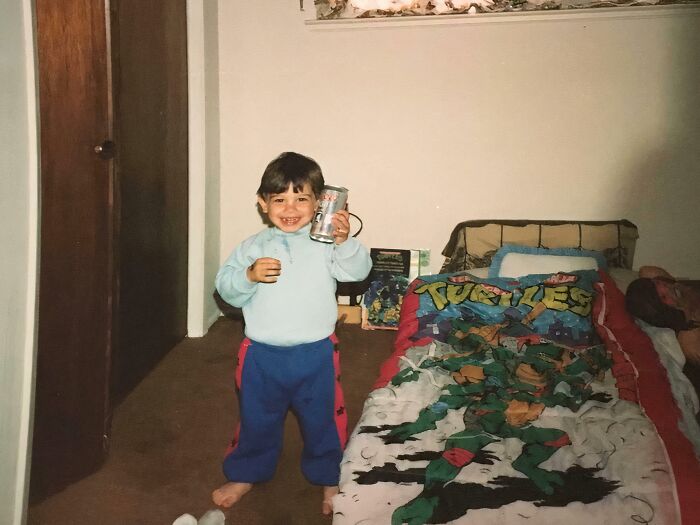 Child holding a drink and smiling in a bedroom with Teenage Mutant Ninja Turtles bedding, capturing an awkward childhood moment.