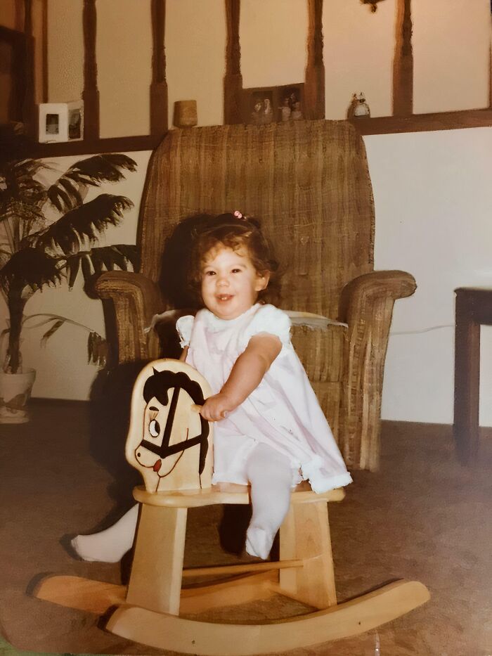 Toddler on a wooden rocking horse in living room, capturing a hilariously awkward childhood pic from the past.