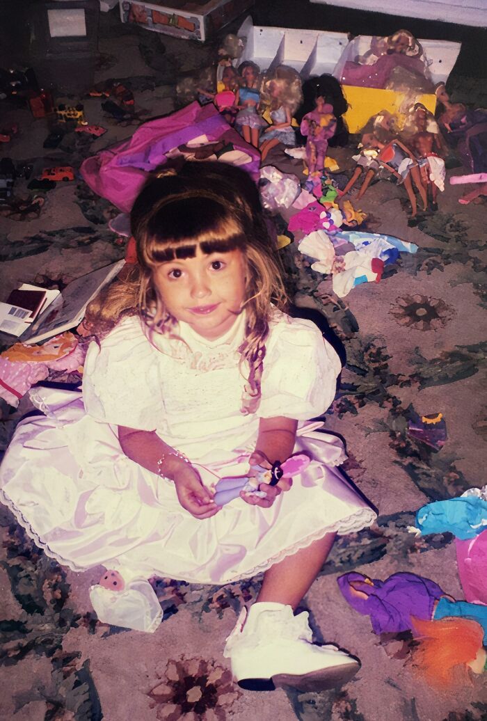 Young girl in a white dress sitting among scattered toys and dolls, a hilariously awkward childhood photo moment.