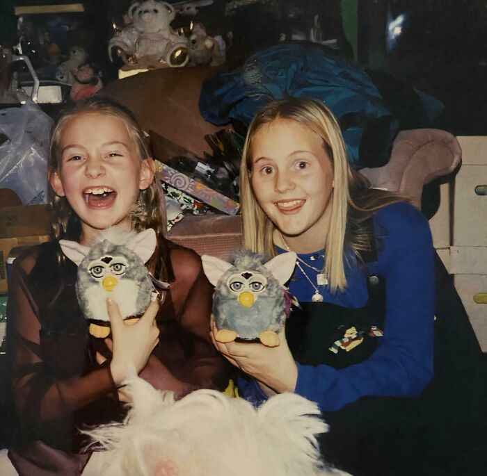 Two girls smiling awkwardly while holding matching Furby toys in a cluttered room, childhood pics with funny moments.