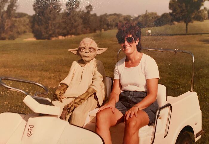 Woman sitting in a golf cart next to a Yoda character, capturing a hilariously awkward childhood pic outdoors.