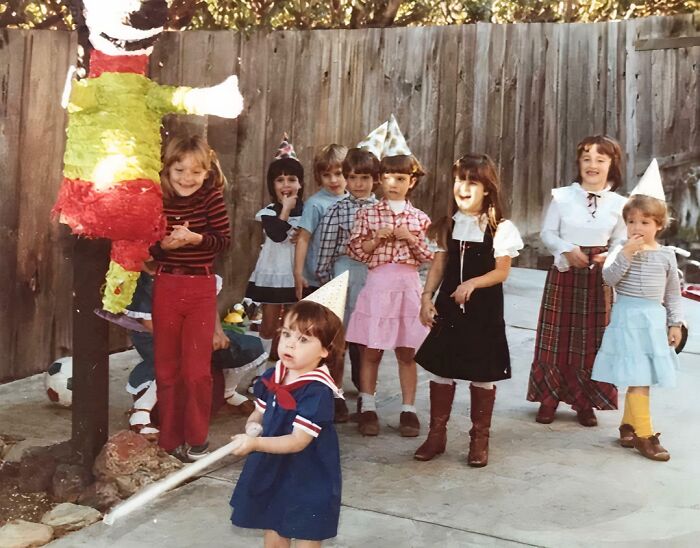 Group of children wearing party hats at a backyard birthday party, one child blindfolded holding a stick near a piñata.