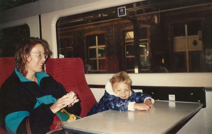 Woman smiling at a child with a mischievous grin sitting at a train table in a hilariously awkward childhood pic.