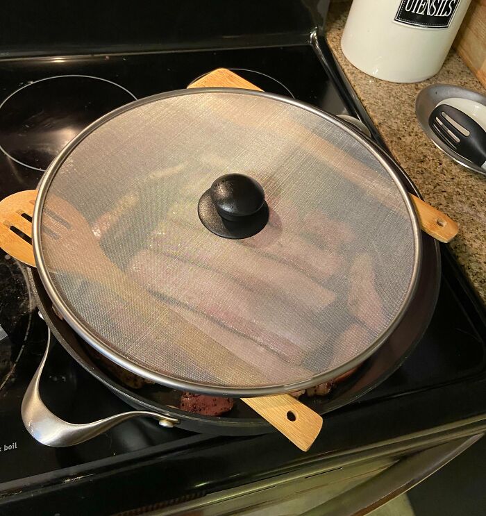 Cooking bacon under a splatter screen held up by wooden spatulas on a stovetop, a genius problem-solving idea.