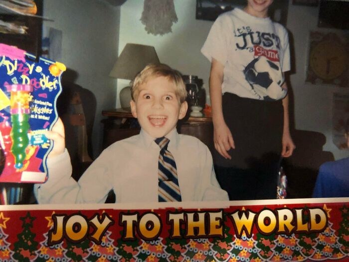 Child holding a toy with a wide smile, standing behind a colorful Joy to the World banner in a humorous childhood photo.