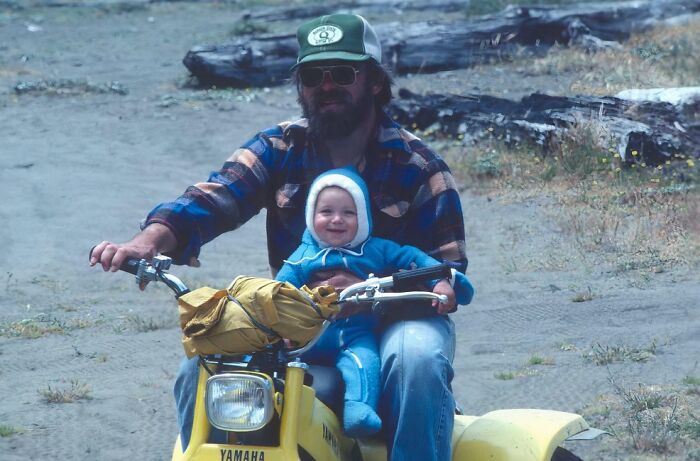 Man with sunglasses and cap rides a yellow Yamaha with a smiling toddler in a blue outfit in a vintage childhood photo.