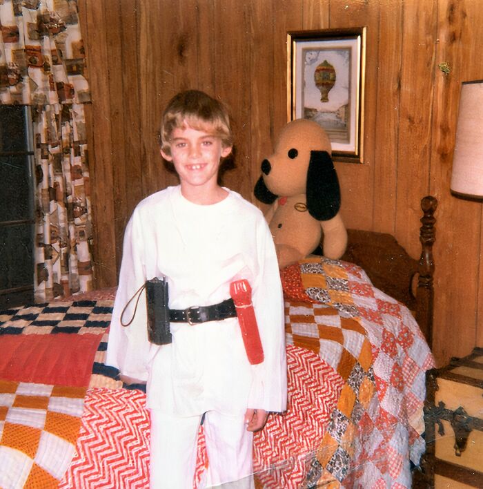 Child in a homemade costume standing in a vintage bedroom with a large stuffed dog and colorful quilt, childhood awkward moment.