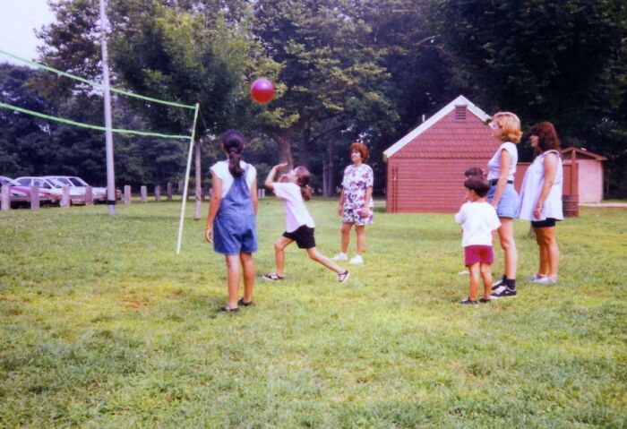 Children playing volleyball in a park, capturing a hilariously awkward childhood moment with family watching nearby.