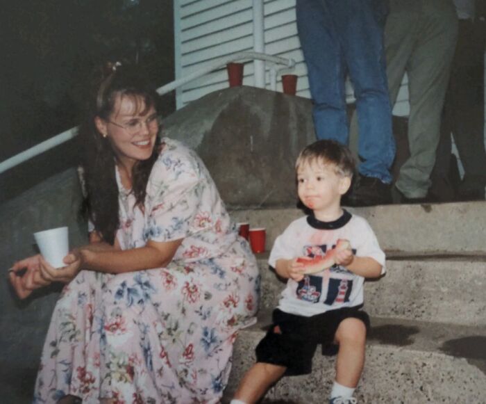 Woman in floral dress smiling at a young boy eating watermelon in a hilariously awkward childhood photo outdoors.