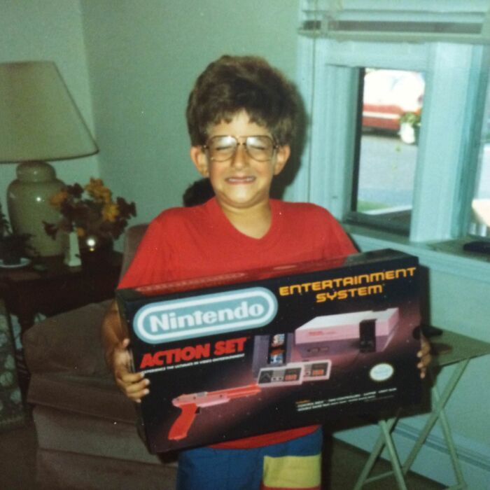 Child holding a vintage Nintendo Entertainment System box, an awkward childhood moment captured indoors.