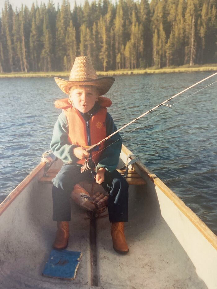 Child fishing in a boat wearing a life jacket and cowboy hat, an awkward childhood pic by the lake with trees in the background.