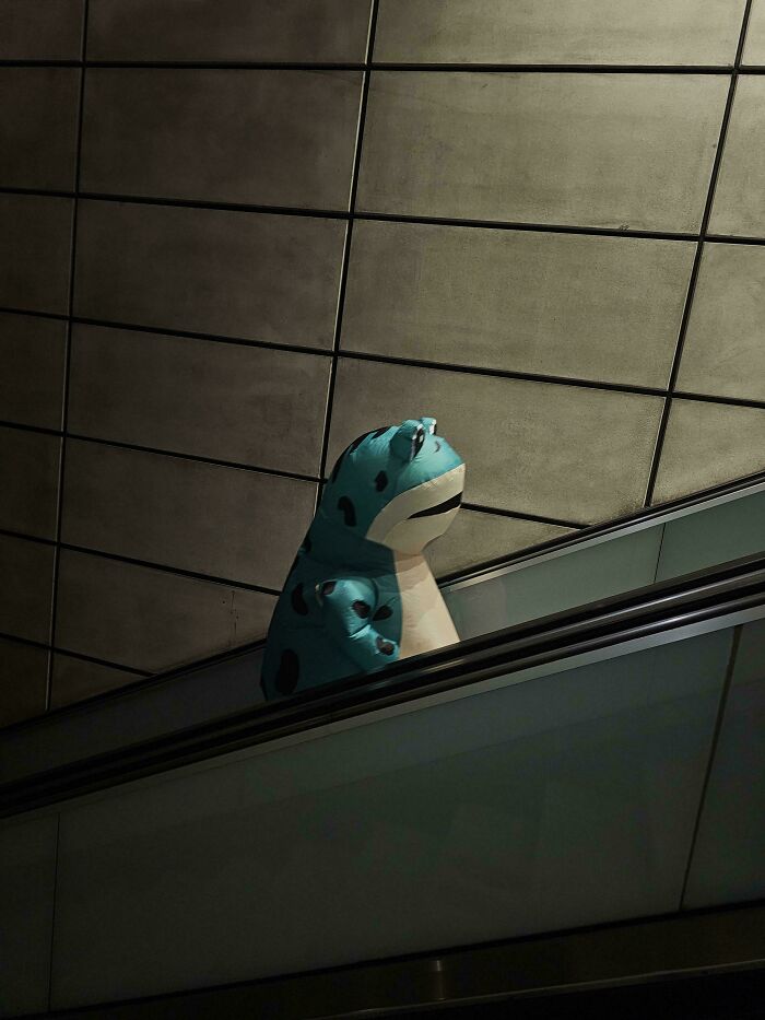 A person in a blue frog costume on an escalator, evoking an Accidental Renaissance style in soft light