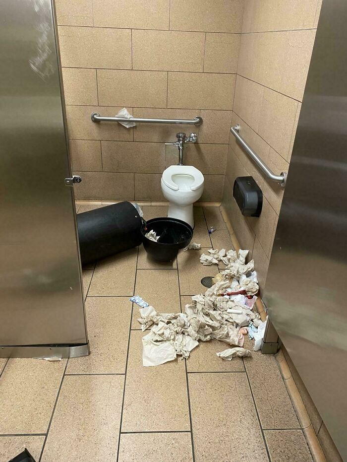 Messy public restroom with trash can tipped over and paper towels scattered on floor showing people operating at 1% brain capacity.