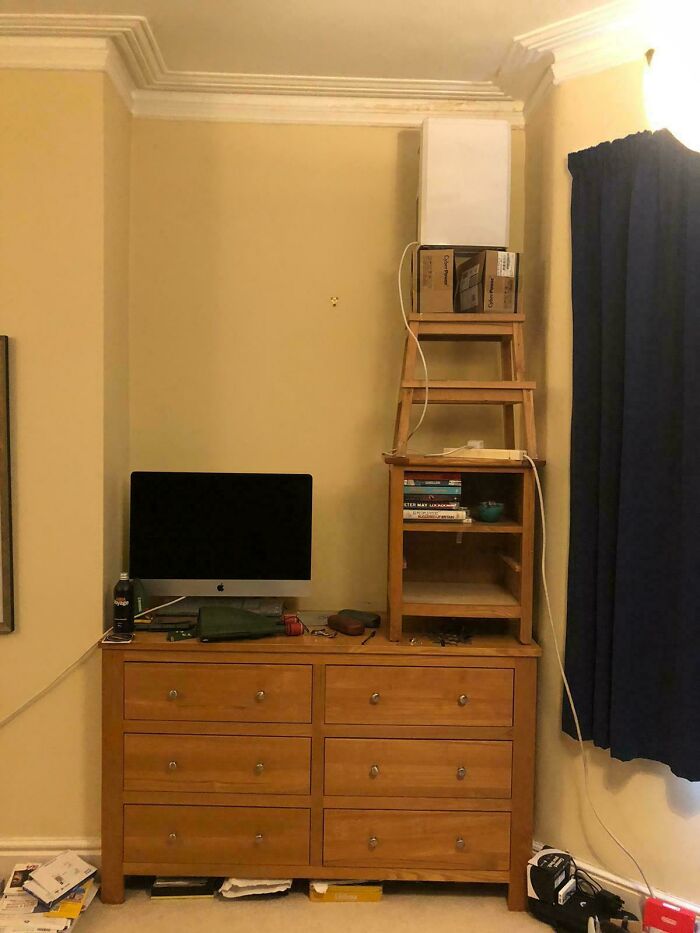 Improvised setup with a router placed on stacked furniture next to a computer on a wooden dresser in a room corner.