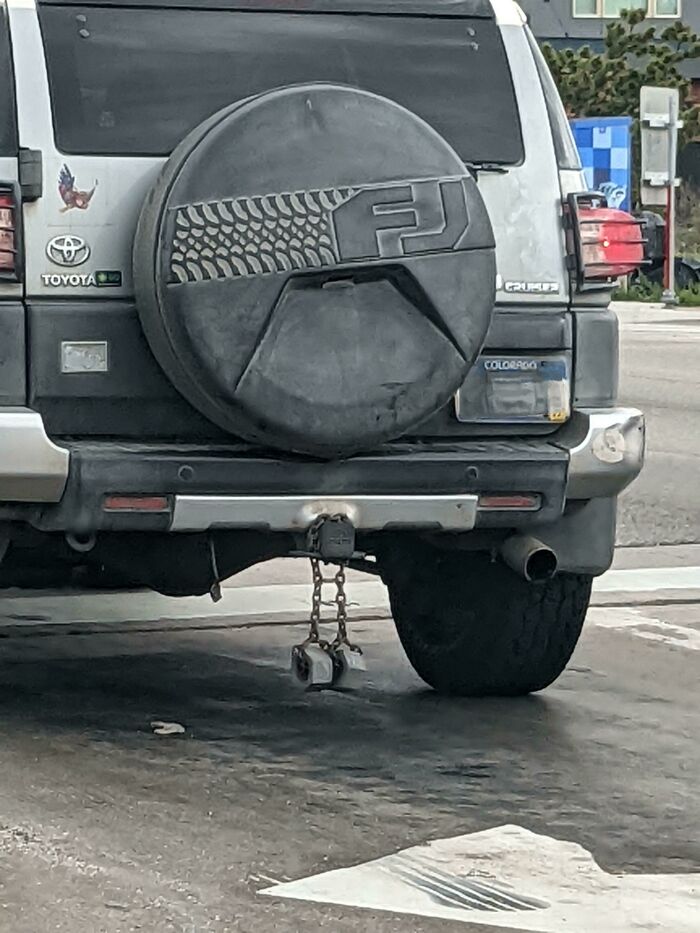 Toyota SUV with a spare tire cover and trailer hitch lock hanging from rusty chains, illustrating people operating at 1% brain capacity.