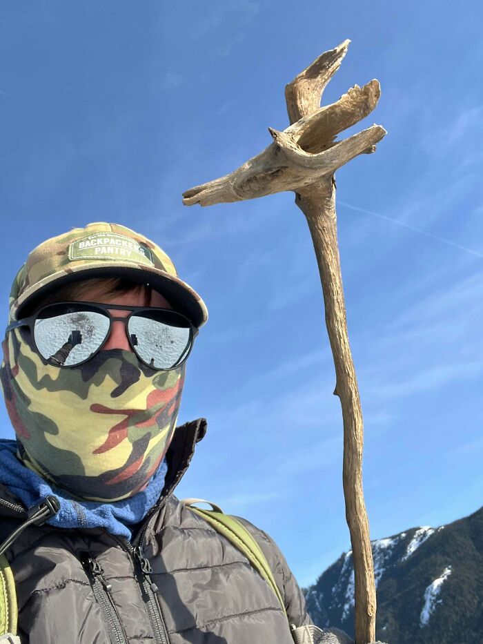 Person wearing camo face covering and hat holding a cool stick outdoors with mountains and clear blue sky background.