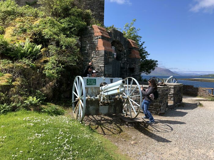 Tourists posing on historic cannon near stone ruins by water under clear blue sky, showcasing rude and entitled behavior.