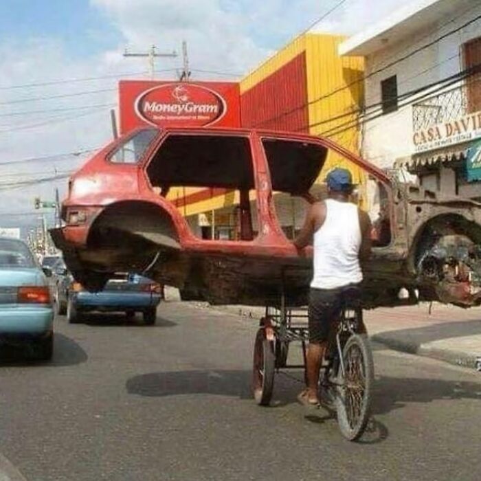 Man on a tricycle carrying a stripped car shell on a busy street in a weird and cringey photo.