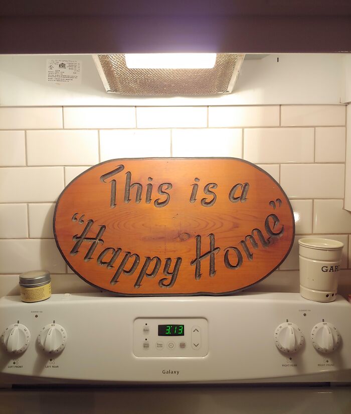 Wooden sign with suspicious quotation marks around the phrase happy home, displayed above a white stove.
