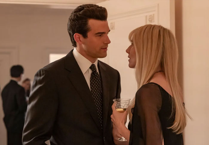 A man in a black suit and a woman with long blonde hair holding a drink in a dimly lit indoor setting.