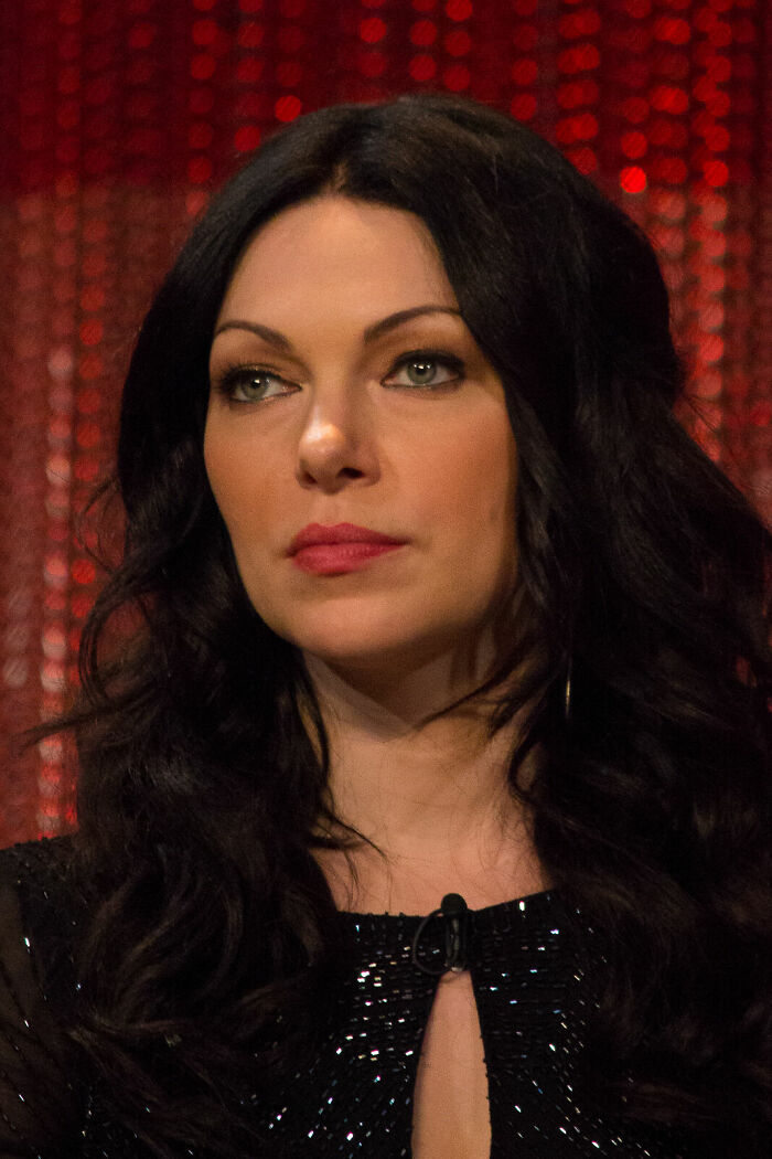 Laura Prepon with long dark hair and subtle makeup, wearing a black outfit in a studio setting with red backdrop.