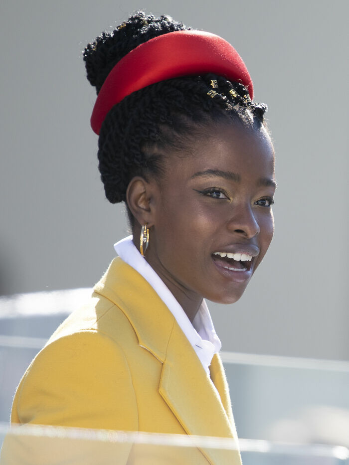 Amanda Gorman wearing a yellow coat and red headband, smiling and speaking outdoors in natural light.
