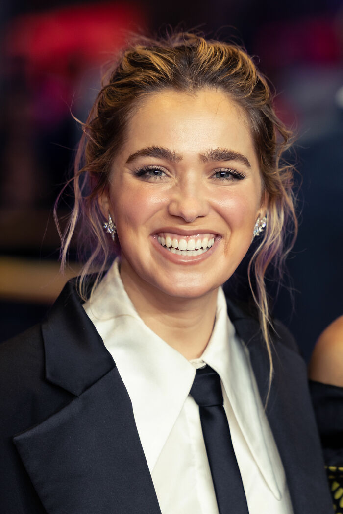 Haley Lu Richardson smiling at an event, wearing a black blazer and tie, highlighting her bio and career highlights.