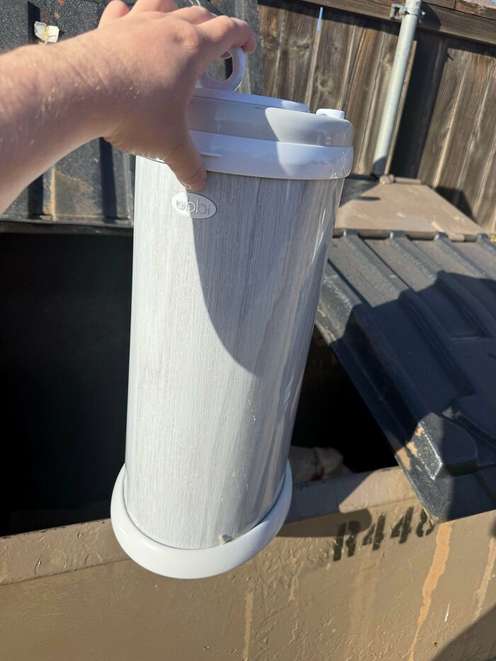 A hand holds a baby diaper pail over a dumpster, representing the funny side of being a great dad.