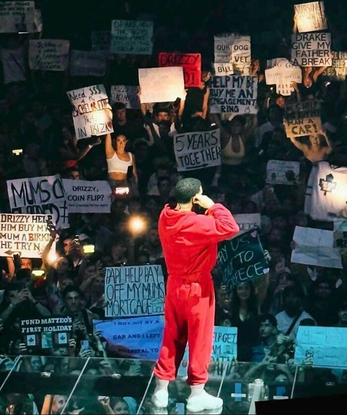 Man in red outfit at concert stage facing crowd holding various protest signs