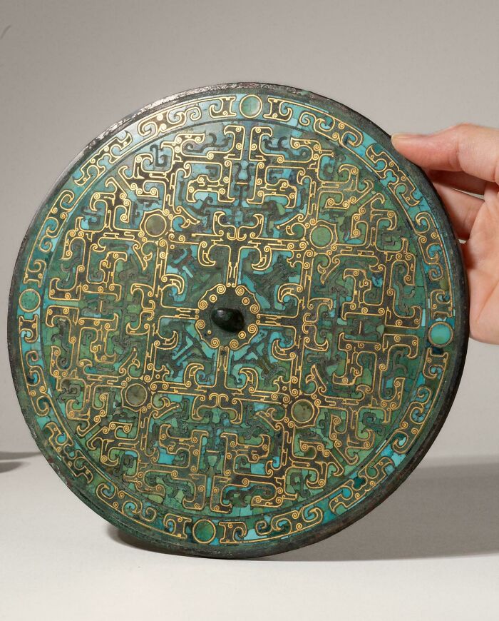 A hand holding an intricately detailed ancient bronze mirror with turquoise inlay from historical finds.