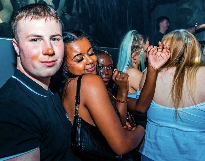 A group of people in a dimly lit club, a man in the foreground with orange paint on his face, peculiar things.