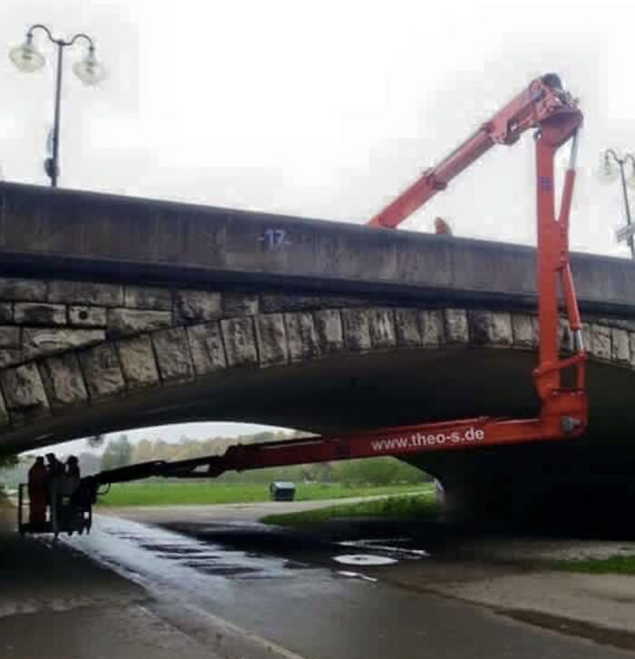 A peculiar sight in real life of a large orange boom lift oddly positioned under and over a stone bridge.