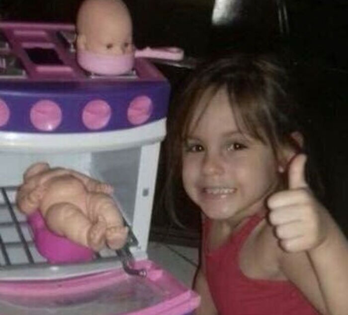A peculiar moment captured in real life of a young girl posing thumbs up with baby dolls in a toy oven and pot.